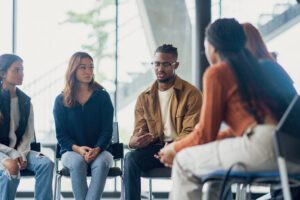 A small group of University students sit together during a group therapy session. They are each dressed casually as they take turns sharing their struggles with the group.