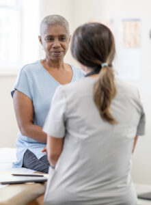 A middle aged woman of African decent, who is using Spravato for treatment, sits up on an exam table for a check-up. She is dressed casually and has a neutral expression o her face. The female nurse is seated in front of her in light grey scrubs as the two talk about how she is feeling and how to combat her side effects.