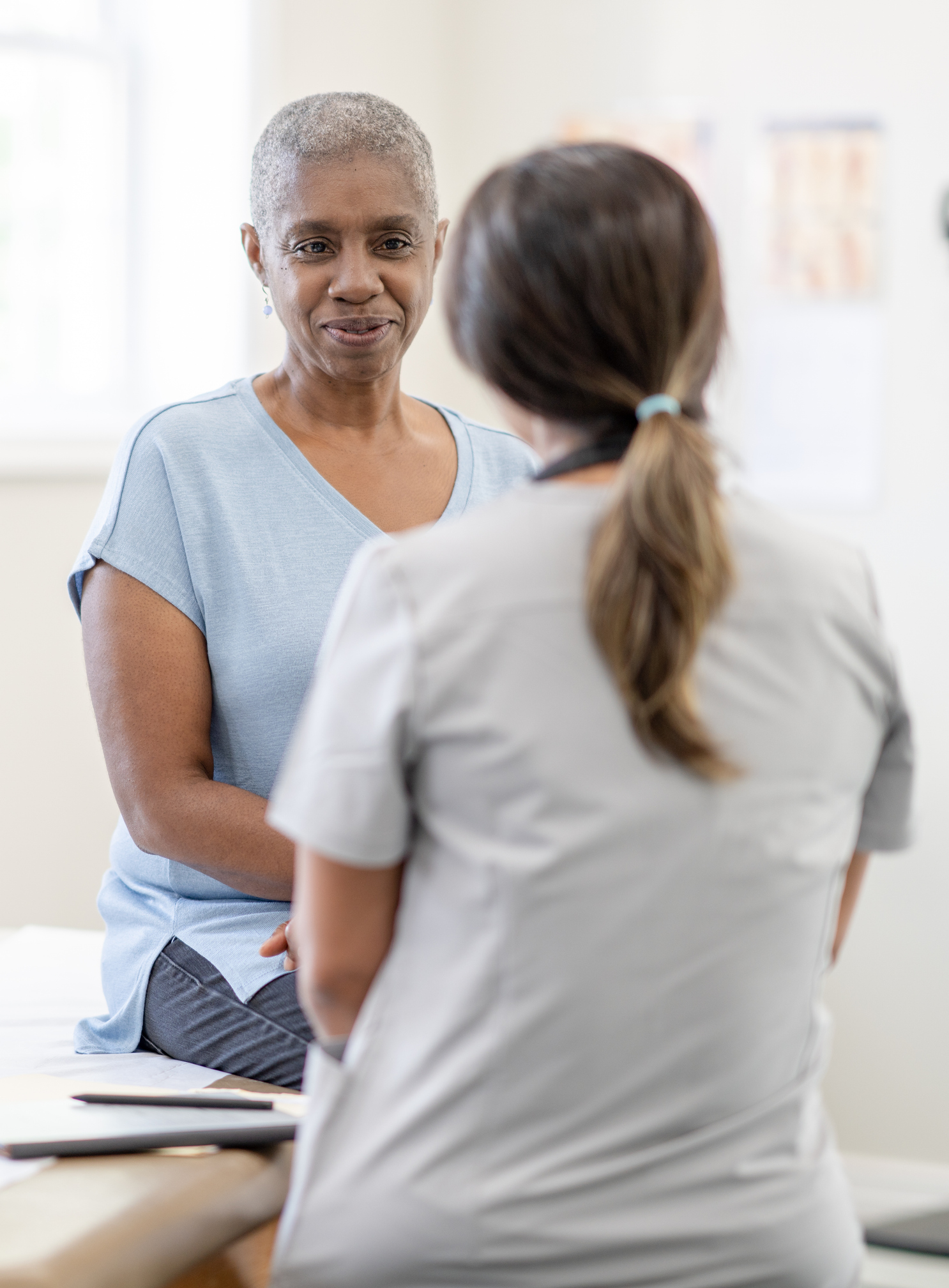A middle aged woman of African decent, who is using Spravato for treatment, sits up on an exam table for a check-up. She is dressed casually and has a neutral expression o her face. The female nurse is seated in front of her in light grey scrubs as the two talk about how she is feeling and how to combat her side effects.