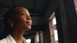 Portrait of a confident Black woman in a modern industrial office setting, gazing thoughtfully. The image captures feelings of ambition and introspection with brick walls and large windows in the background.